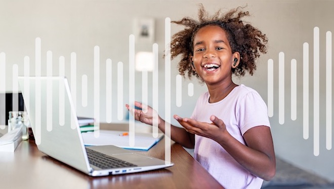 A little girl in front of a laptop, listening through a set of black earbuds, smiling
