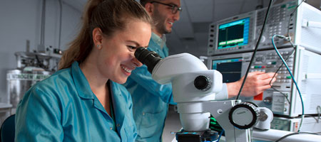 Two engineers smile while examining a device under a microscope.