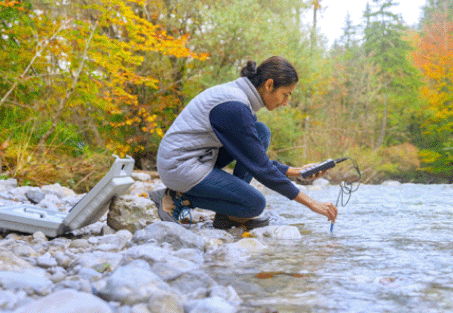 A woman kneeling next to a river and using a device to collect data on the water quality